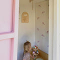 A room with pink walls featuring wall decals of falling flowers, a child standing in front of a door looking at the wall decals.