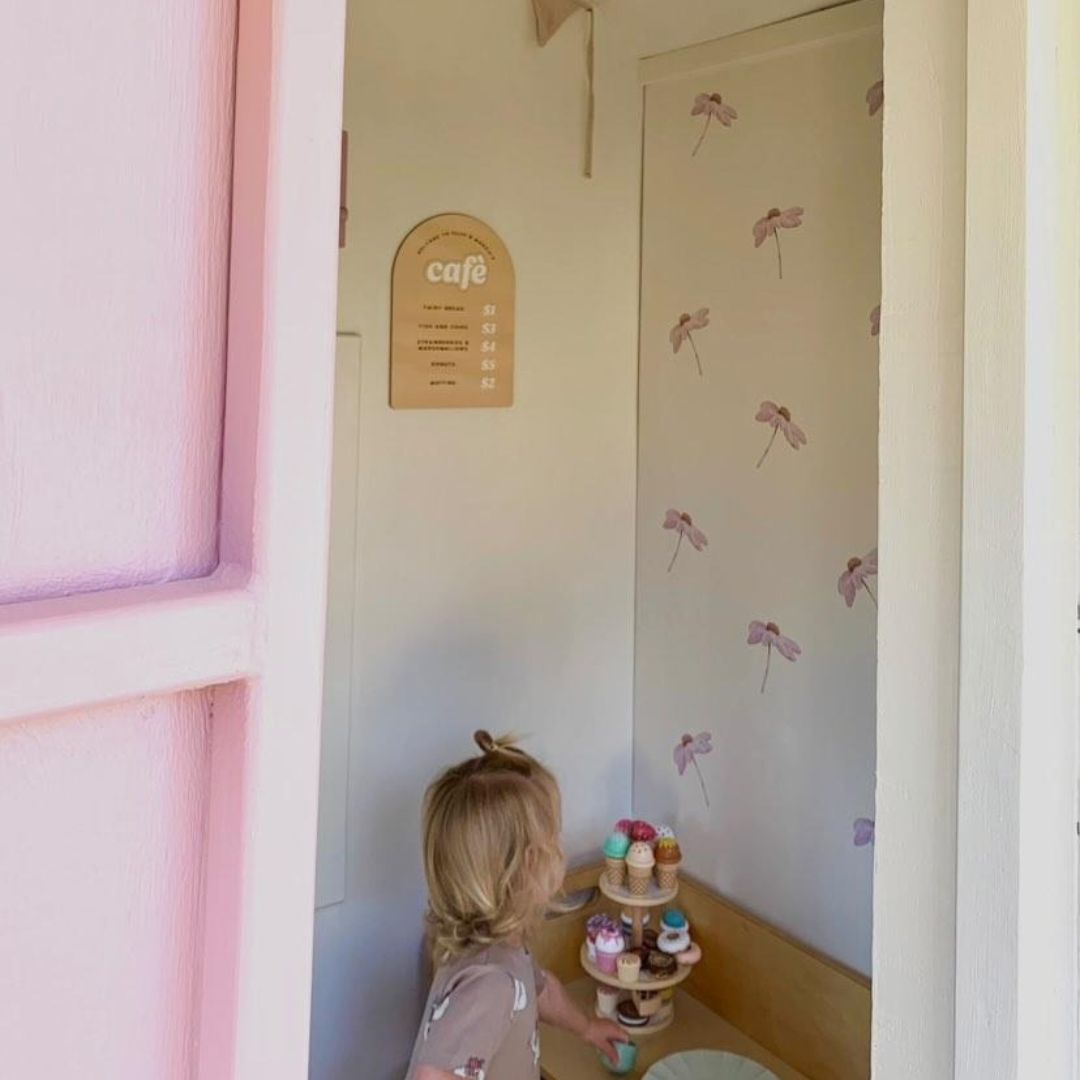 A room with pink walls featuring wall decals of falling flowers, a child standing in front of a door looking at the wall decals.